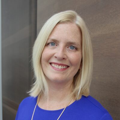 Music therapist, Dr. Abbey Dvorak, who has straight blonde, shoulder-length hair and is wearing a blue blouse standing in front of a brown background