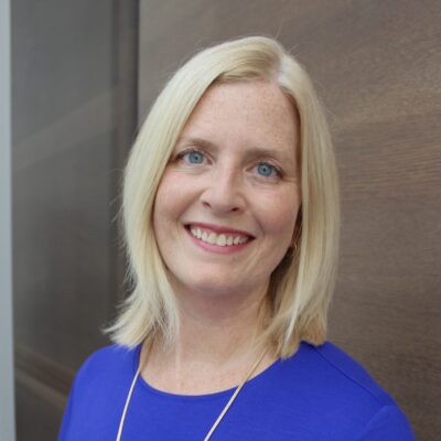 Music therapist, Dr. Abbey Dvorak, who has straight blonde, shoulder-length hair and is wearing a blue blouse standing in front of a brown background