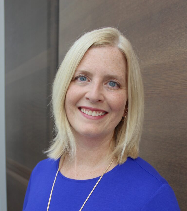 Music therapist, Dr. Abbey Dvorak, who has straight blonde, shoulder-length hair and is wearing a blue blouse standing in front of a brown background