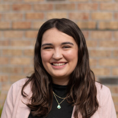 Music therapist, Celia Nowlin, who has long, straight, dark hair and is wearing a black shirt with a light pink sweater, standing in front of a brick wall.