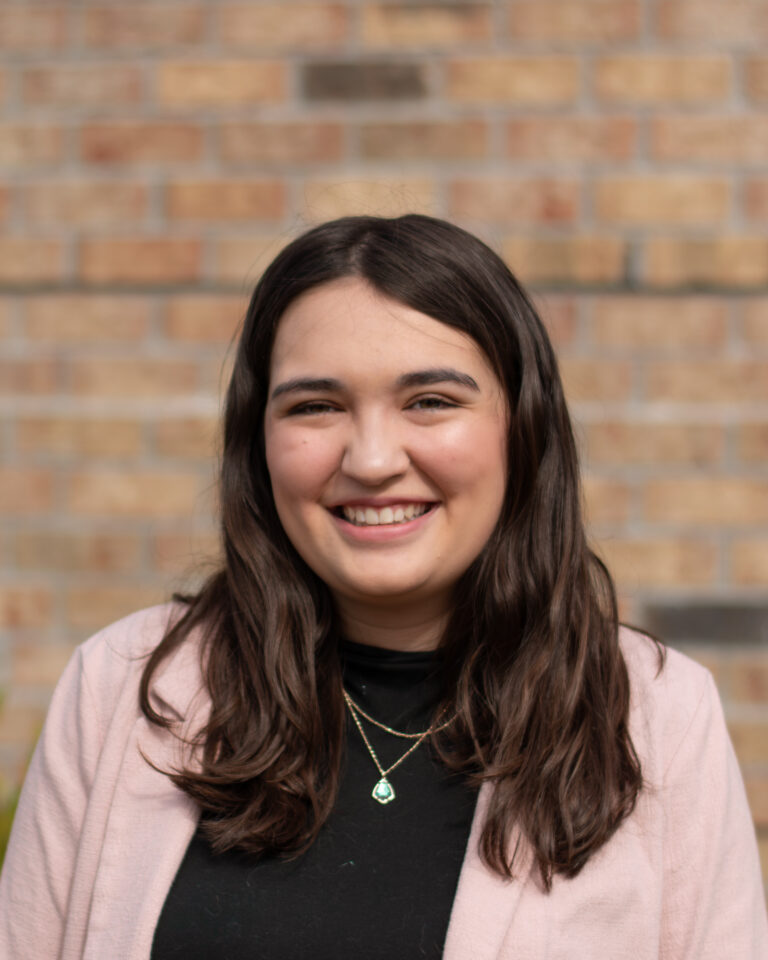 Music therapist, Celia Nowlin, who has long, straight, dark hair and is wearing a black shirt with a light pink sweater, standing in front of a brick wall.