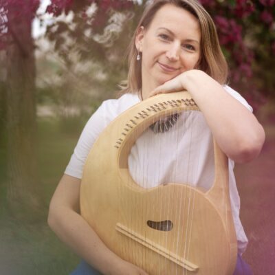 Music therapist, Evgeniya Chernyaeva, who has straight, medium-length, blondish hair, and is standing in front of a tree with dark pink blossoms while holding a stringed instrument.