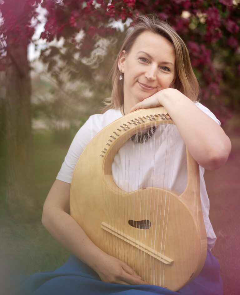 Music therapist, Evgeniya Chernyaeva, who has straight, medium-length, blondish hair, and is standing in front of a tree with dark pink blossoms while holding a stringed instrument.