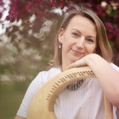 Music therapist, Evgeniya Chernyaeva, who has straight, medium-length, blondish hair, and is standing in front of a tree with dark pink blossoms while holding a stringed instrument.