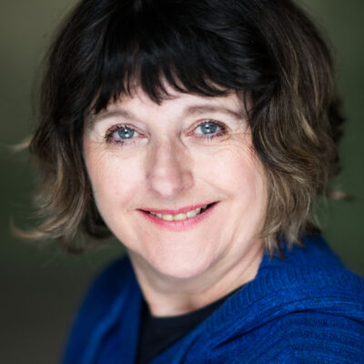 Music therapist, Dr. Elizabeth Coombes, who has short, wavy, brownish-blonde hair. She is wearing a blue sweater in front of a gray background.