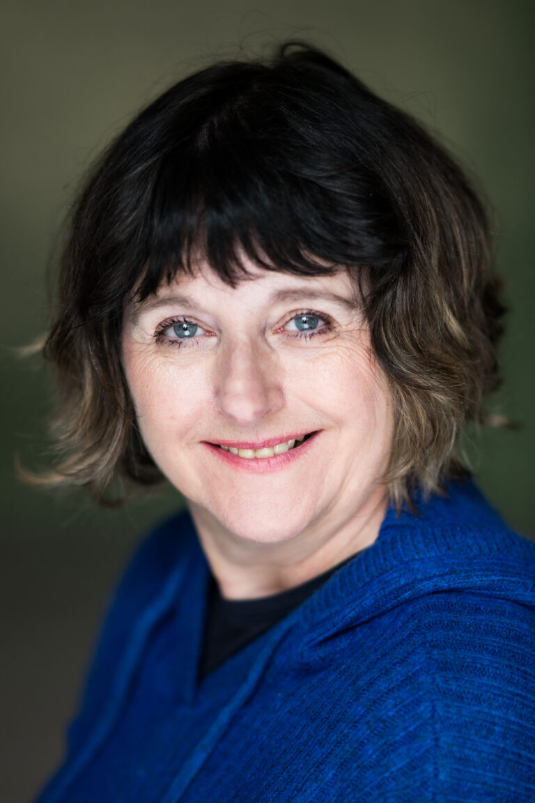 Music therapist, Dr. Elizabeth Coombes, who has short, wavy, brownish-blonde hair. She is wearing a blue sweater in front of a gray background.