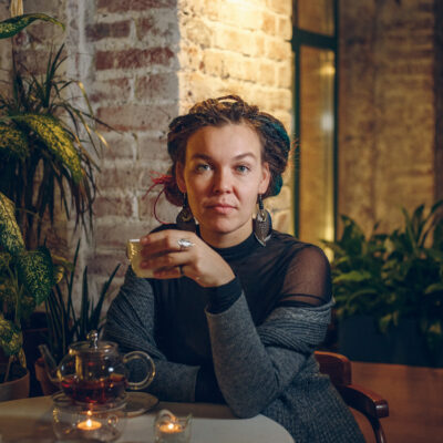 Music therapist, Dr. Ekaterina Molodykh, who is sitting in front of a brick wall with plants surrounding her. She has multi-colored dreadlocks that are pulled back. She is wearing a gray sweater and black top.