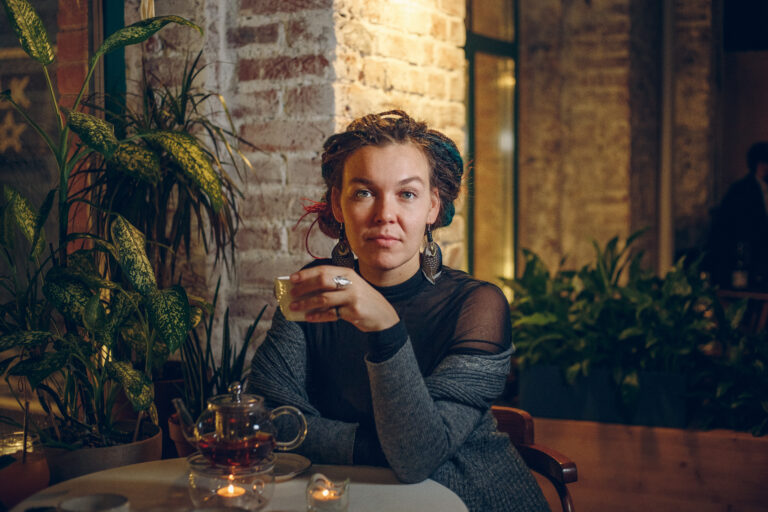 Music therapist, Dr. Ekaterina Molodykh, who is sitting in front of a brick wall with plants surrounding her. She has multi-colored dreadlocks that are pulled back. She is wearing a gray sweater and black top.