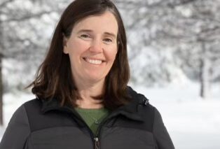 Music therapist, Colleen Klym, who has long, straight, brown hair and is wearing a grey jacket while standing outside in front of a snowy landscape