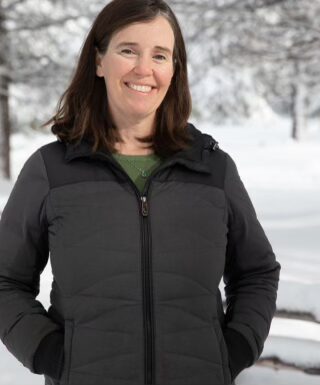 Music therapist, Colleen Klym, who has long, straight, brown hair and is wearing a grey jacket while standing outside in front of a snowy landscape