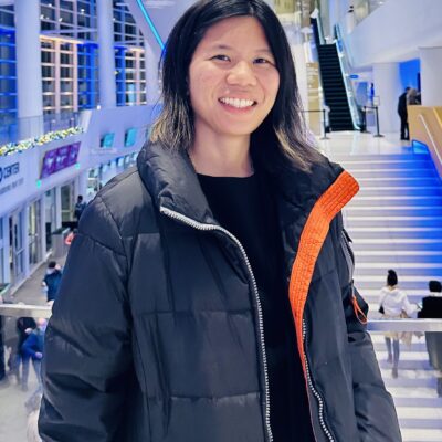 Music therapist, Livia Umeda, who has straight, dark, medium-length hair. She is wearing a black shirt and a navy blue jacket in front of a white and blue background