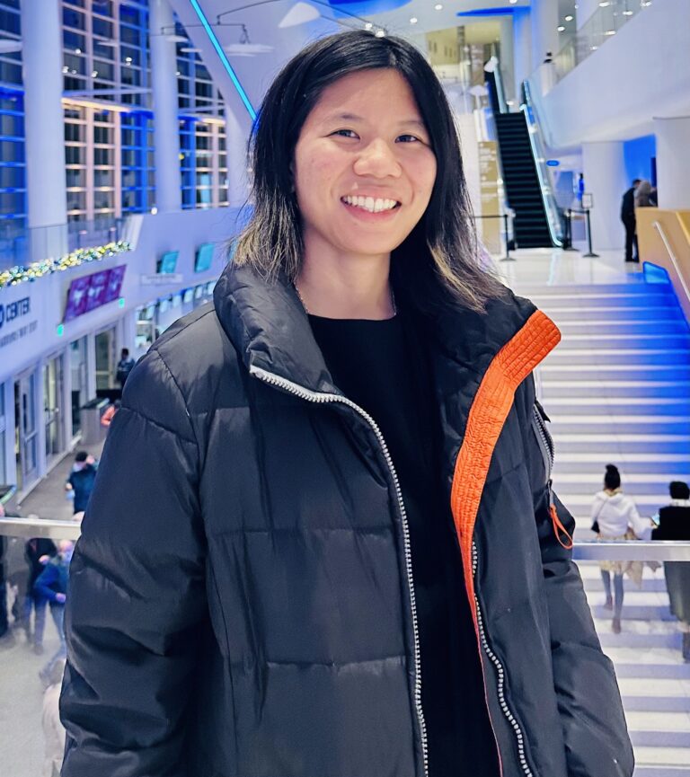 Music therapist, Livia Umeda, who has straight, dark, medium-length hair. She is wearing a black shirt and a navy blue jacket in front of a white and blue background