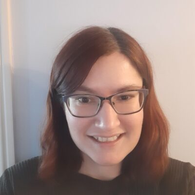 Music therapist, Vicky Guise, who has straight, red hair that is medium length, and is wearing glasses and a brown shirt in front of a white wall.